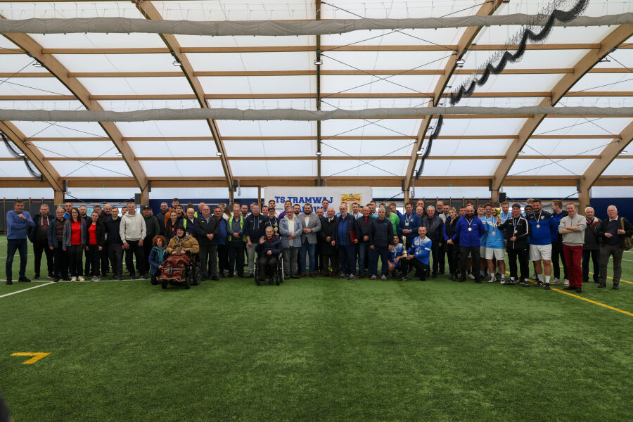 Grupa około 70 osób pozuje do wspólnego zdjęcia na hali sportowej TS Tramwaj. Na pierwszym planie widoczne są osoby na wózkach. W tle baner z nazwą klubu. Zdjęcie zrobione na sztucznej murawie pod zadaszeniem z drewna i tworzywa.