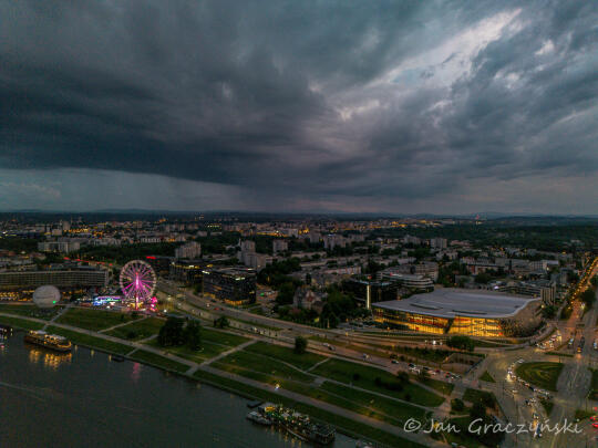 Zdjęcie miasta o zmierzchu pokazuje oświetloną panoramę Krakowa z widocznym kołem widokowym, budynkiem ICE Kraków i rzeką Wisłą na pierwszym planie. Nad miastem wiszą ciemne burzowe chmury.