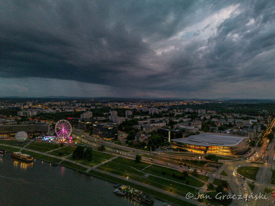 Zdjęcie miasta o zmierzchu pokazuje oświetloną panoramę Krakowa z widocznym kołem widokowym, budynkiem ICE Kraków i rzeką Wisłą na pierwszym planie. Nad miastem wiszą ciemne burzowe chmury.