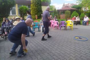 Spotkanie integracyjne przy grillu 30.04.2025 Grupa osób bierze udział w zabawie na świeżym powietrzu; uczestnicy rzucają woreczkami do celu przy dopingu widowni.
