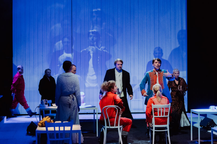 Scena teatralna z kilkoma aktorami w barwnych kostiumach. Tło to jasna ściana z projekcją sylwetek. Postacie stoją i siedzą przy białych krzesłach i stołach, światło jest niebieskawe, tworząc nowoczes