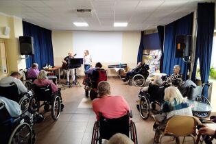 Zajęcia muzyczne 22.05.2025 Kilkunastu mieszkańców słucha dwójki terapeutów śpiewających i grających na gitarze.