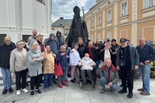 Wycieczka do Wadowic 09.05.2025 Grupa osób pozuje przed pomnikiem, w tle zabytkowe budynki oraz kwietniki.