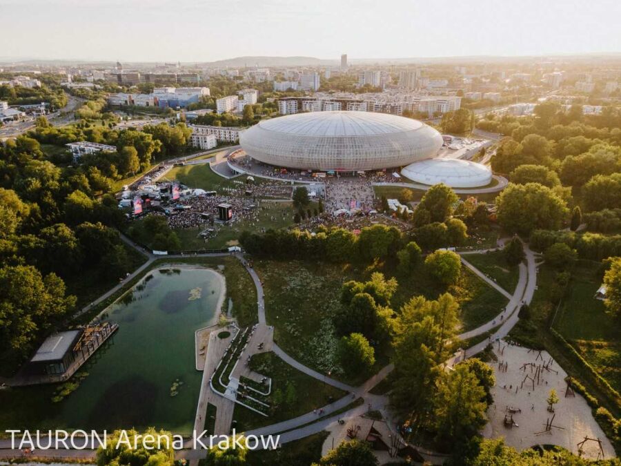 Zdjęcie z drona z TAURON Arena Kraków oraz przylegającymi łąkami, parkiem, zalewem i budynkami mieszkalnymi.
