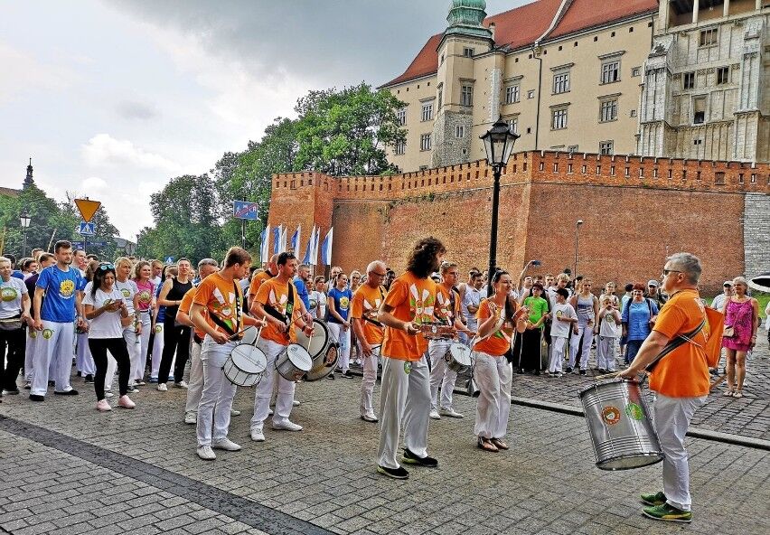 Inauguracja Festiwalu CRACOVIAXE 2025. Na fotografii grupa osób w pomarańczowych strojach z bębnami. W tle zamek Królewski na wawelu