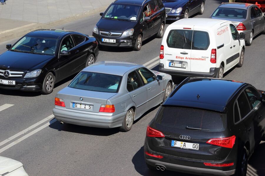 Ruch drogowy w mieście. Na zdjęciu widoczna dwupasmowa jezdnia i kilkanaście aut różnych marek z niemieckimi nr rejestracyjnymi w ruchu ulicznym. W górnym lewym rogu widoczny szeroki chodnik.