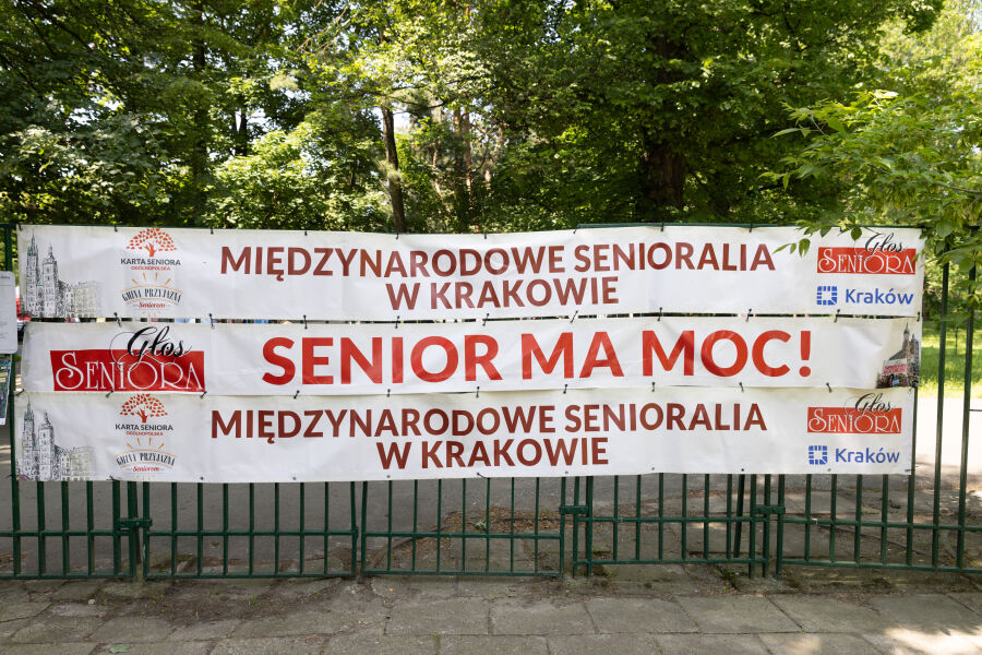 Zdjęcie zawiera baner z XII Międzynarodowych Senioraliów w Krakowie zawieszony na płocie.