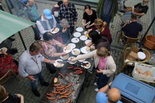 Dzień Rodziny 03.06.2025 Grupa osób zebrana przy dużym grillu z kiełbaskami na świeżym powietrzu. Przygotowywany jest posiłek, rozkładane są talerze z chlebem, musztardą i keczupem. Część osób siedzi przy stolikach.