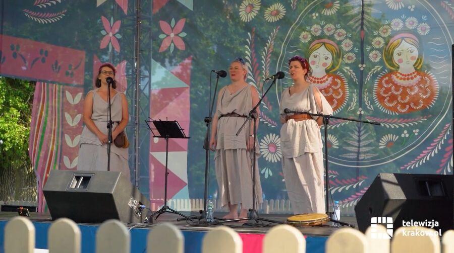 Na scenie trzy kobiety w jasnych strojach ludowych śpiewają do mikrofonów. W tle kolorowa dekoracja z motywami kwiatowymi i postaciami. Na dole logo „telewizja krakow.pl”. Występ odbywa się na zewnątr