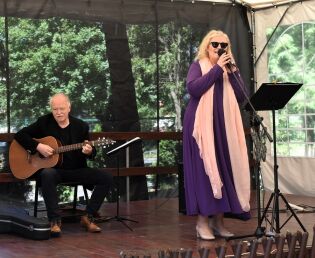 Na zdjęciu widzimy występ muzyczny na scenie plenerowej. Po lewej stronie siedzi  mężczyzna grający na gitarze akustycznej. Jest ubrany na czarno i skupiony na grze. 