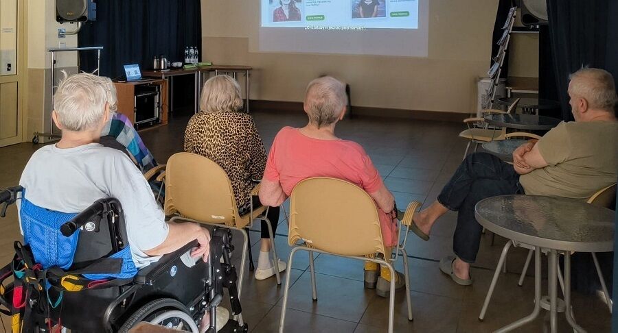 Na zdjęciu widoczna jest grupa osób oglądająca film w sali projekcyjnej. Część widzów siedzi na krzesłach, a także  na wózkach inwalidzkich. 