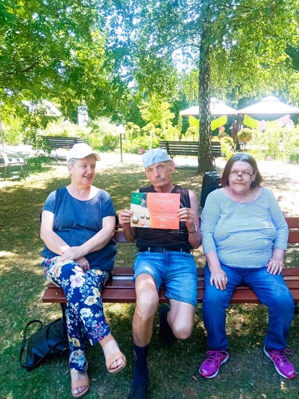 Trzy osoby siedzące na ławce w ogrodzie – mężczyzna w środku trzyma dyplom za udział w turnieju. Towarzyszą mu dwie kobiety, wszyscy uśmiechnięci, w tle zielone drzewa, parasole i ławki.