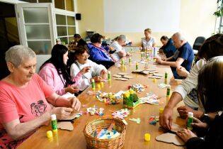 grupa osób w różnym wieku, biorących udział w warsztatach plastycznych. Uczestnicy siedzą przy dużym stole, na którym rozłożone są kolorowe elementy wycięte z papieru, kartonowe kształty.