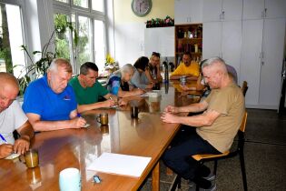 Grupa  osób siedzi przy dużym stole w jasnym pomieszczeniu z dużymi oknami. Każda osoba jest pochylona nad kartką papieru, pisząc coś długopisem.