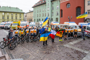 Kraków powitał uczestników rowerowej akcji charytatywnej Bike Convoy for Ukraine W centrum płyty Małego Runku stoją od lewej Konsul Generalny Ukrainy z flagą narodową, Sekretarz Miasta Krakowa z polską flagą i Konsul Generalny Niemiec z niemiecką flagą za nim rowerzyści i karetki