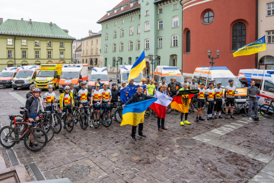 W centrum płyty Małego Runku stoją od lewej Konsul Generalny Ukrainy z flagą narodową, Sekretarz Miasta Krakowa z polską flagą i Konsul Generalny Niemiec z niemiecką flagą za nim rowerzyści i karetki