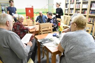Grupa  osób siedzi przy drewnianych stołach w bibliotece. Na stołach stoją wazony z kolorowymi kwiatami, a uczestnicy trzymają w rękach kartki. 