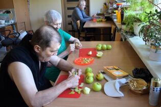 Kulinarne 14.07.2025 Trzy osoby siedzą przy stołach w jasnym pomieszczeniu i obierają zielone jabłka.