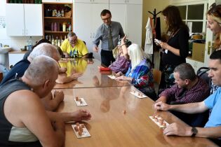 Turniej lotto 14.07.2025 Grupa mieszkańców Domu Pomocy Społecznej siedzi wokół dużego stołu. Każdy uczestnik ma przed sobą planszę z numerkami.