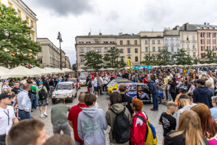 Wielki finał Wielkiej Wyprawy Maluchów na Rynku Głównym Zdjęcie z tłumem ludzi zgromadzonych wokół Fiatów 126P na Rynku Głównym w Krakowie. W tle kamienice, drzewa, kawiarniane. Uczestnicy patrzą na auta, robią zdjęcia. Scena plenerowa, pochmurne niebo.