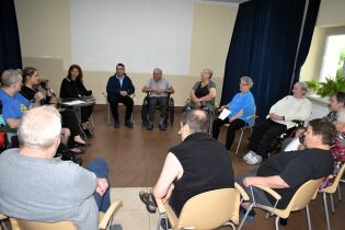 Spotkanie ze sceną 16.07.2025 Grupa starszych osób siedzących w kręgu podczas spotkania w sali. Część uczestników korzysta z wózków inwalidzkich. Jedna z kobiet mówi do mikrofonu, a inna osoba robi notatki. W tle jasna ściana.