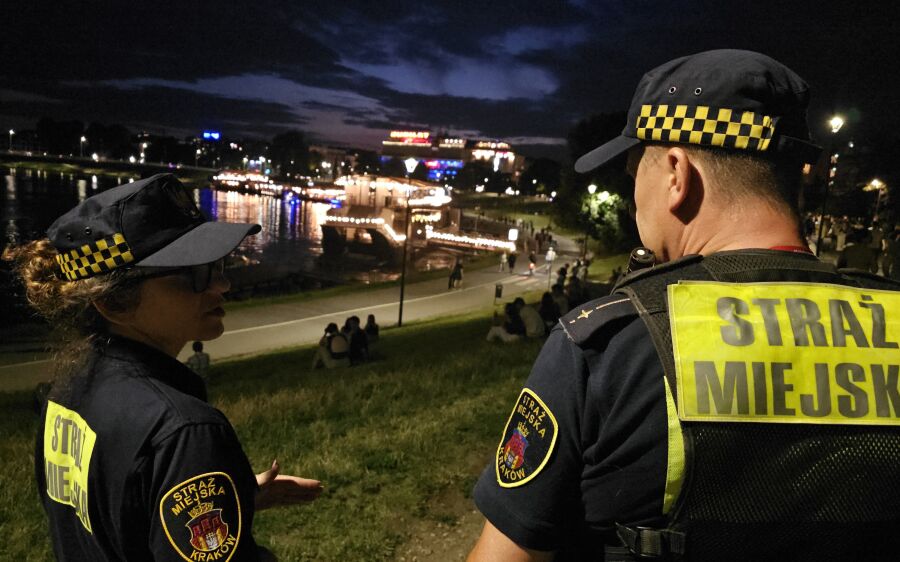 Na zdjęciu dwójka strażników miejskich patroluje nocą okolice Bulwarów Wiślanych w Krakowie. W tle widać Wisłę, oświetlone barki oraz spacerujących ludzi. Zdjęcie wykonane po zmroku.