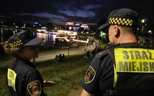 Na zdjęciu dwójka strażników miejskich patroluje nocą okolice Bulwarów Wiślanych w Krakowie. W tle widać Wisłę, oświetlone barki oraz spacerujących ludzi. Zdjęcie wykonane po zmroku.