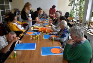 Arteterapia 18.07.2025 Uczestnicy, w większości seniorzy i osoby z niepełnosprawnościami, wykonują prace plastyczne z kolorowego papieru i kleju. Nad przebiegiem zajęć czuwają opiekunowie.