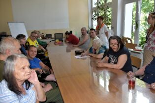 Kalambury i gry słowne 21.07.2025 Grupa dorosłych osób siedzi przy długim stole w jasnym pomieszczeniu. Uczestnicy są zaangażowani w zajęcia integracyjne. W tle stoją opiekunki.