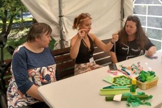 Warsztaty makramy 22.07.2025 Trzy kobiety siedzą przy stole na zewnątrz, w namiocie ogrodowym. Na stole leżą kolorowe materiały, wstążki, papier, klej i gotowe ozdoby. Kobieta po środku pokazuje coś palcami, jakby instruowała