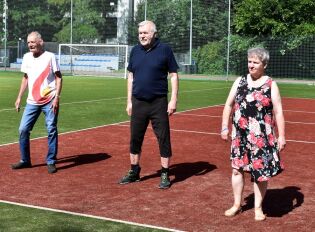 Festiwal Tańców Dworskich 23.07.2025 Trzy osoby – dwaj mężczyźni i jedna kobieta – stoją na boisku sportowym, częściowo na czerwonej nawierzchni, częściowo na zielonej trawie, uczestniczą w zajęciach ruchowych na świeżym powietrzu.