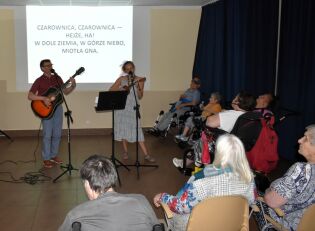 Karaoke 24.07.2025 Grupa osób, głównie seniorów i osób z niepełnosprawnościami, siedzi i słucha występu duetu muzycznego – mężczyzna z gitarą i kobieta z ukulele śpiewają na scenie. Na ścianie za nimi wyświetlony jest f
