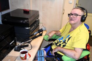Audycja radiowęzła 29.07.2025 Na zdjęciu widzimy mężczyznę siedzącego na wózku inwalidzkim, który prowadzi audycję radiową. Ma na sobie żółtą koszulkę i słuchawki na uszach. Przed nim na biurku znajduje się mikrofon na statywie.