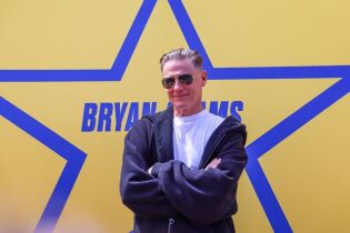 Bryan Adams w okularach przeciwsłonecznych, białym t-shircie i czarnej bluzie na tle żółtej planszy z gwiazdą z niebieskim konturem  i jego nazwiskiem w jej centrum. 