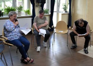 Spotkanie ze sceną 30.07.2025 Trzy osoby siedzą w kręgu w jasnym pomieszczeniu z dużymi oknami i roślinami doniczkowymi. Kobieta z mikrofonem czyta tekst z kartki, druga kobieta i mężczyzna obok niej również trzymają kartki.