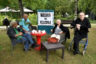 Turniej Mölkky 31.07.2025 Cztery starsze osoby siedzą na zewnątrz przy czerwonym stoliku z filiżankami i talerzykiem po ciastkach. Za nimi wisi kolorowy baner z napisem „MÖLKKY na KLUZEKA”.