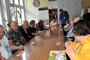 Grupa osób siedzi wokół stołu i gra w grę planszową Lotto. Na stole plansze z liczbami i drewniane baryłki.