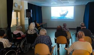 Grupa seniorów siedzi na krzesłach i wózkach inwalidzkich w ciemnej sali, oglądając film z żaglowcem wyświetlany na dużym ekranie.