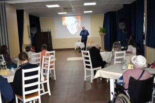 Mężczyzna przemawia przed publicznością w sali, za nim wyświetlone zdjęcie starszej kobiety – Krystyny Miłobędzkiej. Uczestnicy siedzą przy stolikach, słuchając prezentacji.