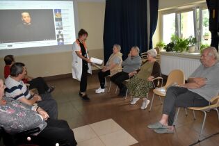 Grupa starszych osób siedzi w półokręgu w sali, oglądając projekcję wideo na ścianie. Przed nimi stoi kobieta w białym fartuchu, prowadząca zajęcia. Za nią na ekranie wyświetlany jest film z YouTube. 