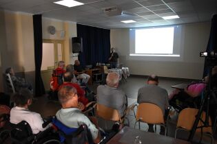 Na zdjęciu widać grupę osób siedzących w sali, skierowanych w stronę ekranu projekcyjnego. Na ekranie wyświetlany jest slajd, a przy nim stoi mężczyzna prowadzący prezentację i mówiący do mikrofonu. 