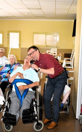 mężczyzna w okularach trzymający mikrofon i pochylający się w stronę mężczyzny na wózku inwalidzkim. Obaj uśmiechają się szeroko, sprawiając wrażenie, że wspólnie śpiewają. 