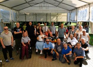 Grupa mieszkańców i pracowników Domu Pomocy Społecznej oraz zaproszonych gości stoi i siedzi wspólnie pod dużym białym namiotem ogrodowym. W centrum widać księdza oraz osoby na wózkach inwalidzkich. 