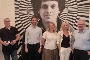 Wizyta studyjna przedstawicieli Muzeum Fotografii w Ołomuńcu. Na zdjęciu trzech mężczyzn i dwie kobiety stoją przodem do patrzącego pod graficznym dziełem sztuki zawieszonym na ścianie muzeum.  