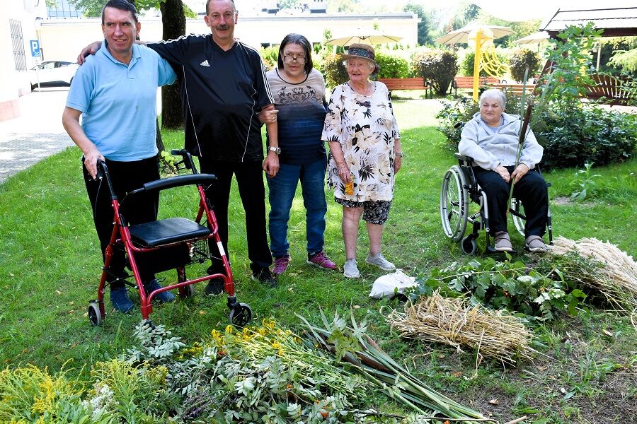 pięć osób stojących i siedzących na trawie w ogrodzie, obok zebranych roślin i ziół. Dwie osoby stoją z lewej strony — jedna z nich opiera się o czerwony balkonik.