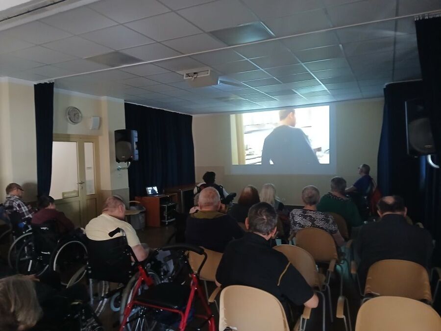 grupa osób siedzących na krzesłach i w wózkach inwalidzkich w ciemniejszej sali, gdzie odbywa się projekcja filmowa. Na ścianie przed nimi wyświetlany jest obraz z projektora.