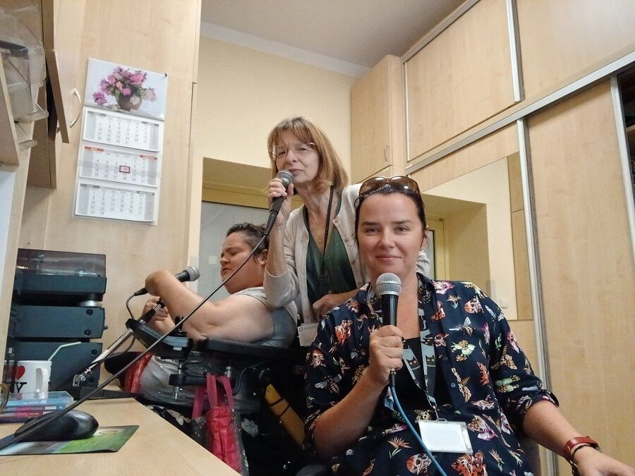 trzy kobiety siedzące w pomieszczeniu radiowęzła. Każda z nich trzyma w ręku mikrofon i uśmiecha się w stronę aparatu. W tle znajdują się drewniane szafki, kalendarz na ścianie oraz sprzęt radiowy.