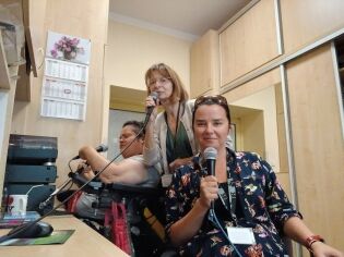 trzy kobiety siedzące w pomieszczeniu radiowęzła. Każda z nich trzyma w ręku mikrofon i uśmiecha się w stronę aparatu. W tle znajdują się drewniane szafki, kalendarz na ścianie oraz sprzęt radiowy.