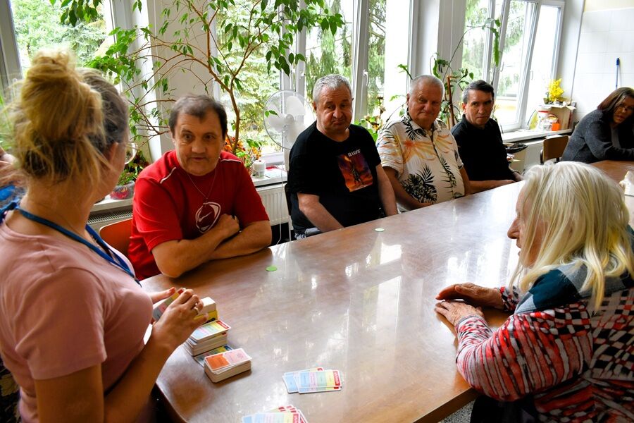 Na zdjęciu widzimy grupę starszych osób siedzących wokół stołu w jasnym, przestronnym pomieszczeniu z dużymi oknami i roślinami doniczkowymi w tle.