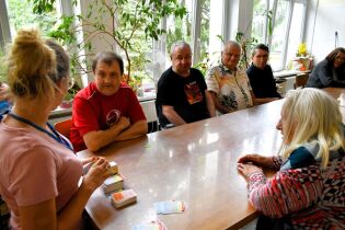 Na zdjęciu widzimy grupę starszych osób siedzących wokół stołu w jasnym, przestronnym pomieszczeniu z dużymi oknami i roślinami doniczkowymi w tle. 